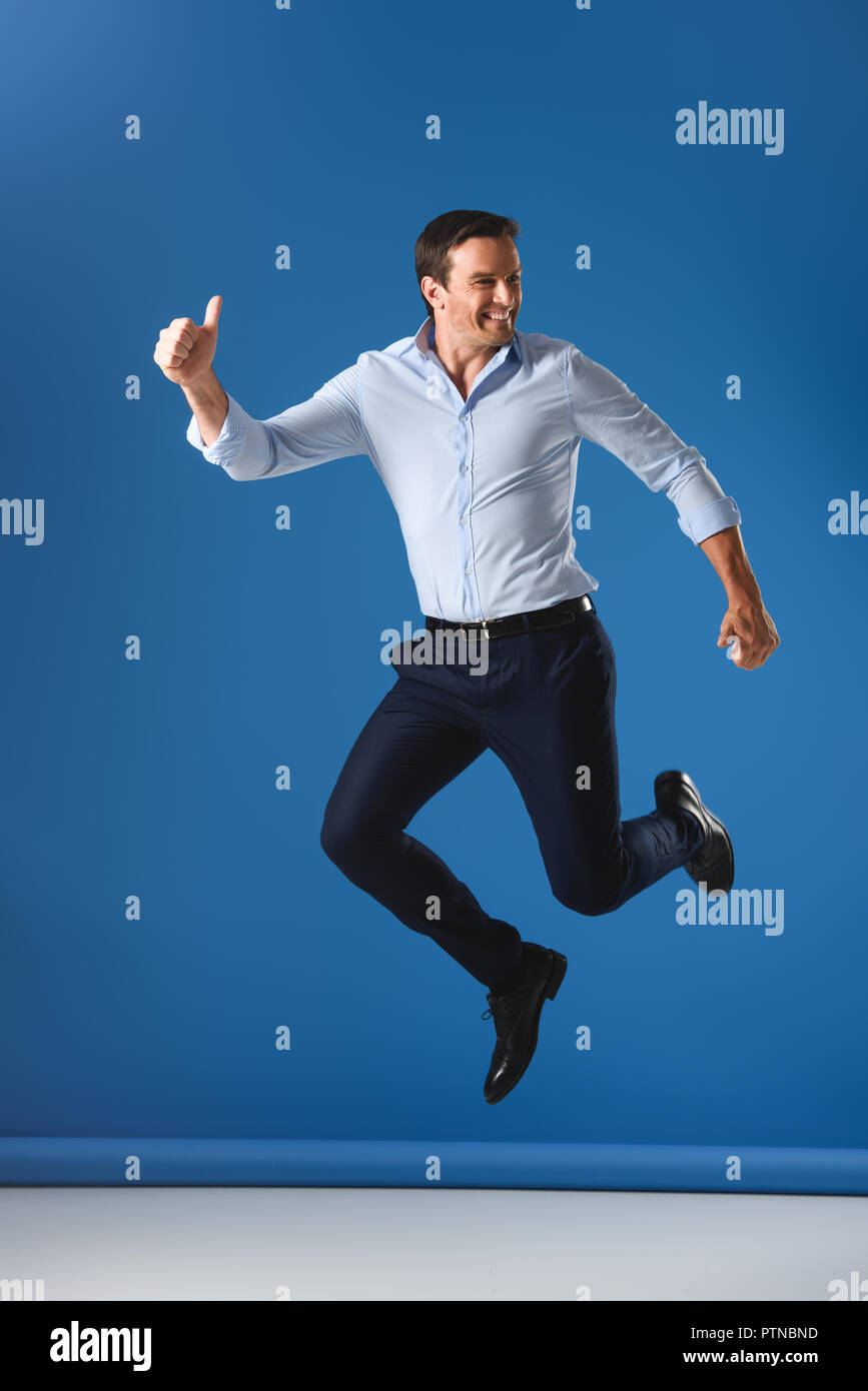 cheerful handsome man showing thumbs up and jumping on blue Stock Photo