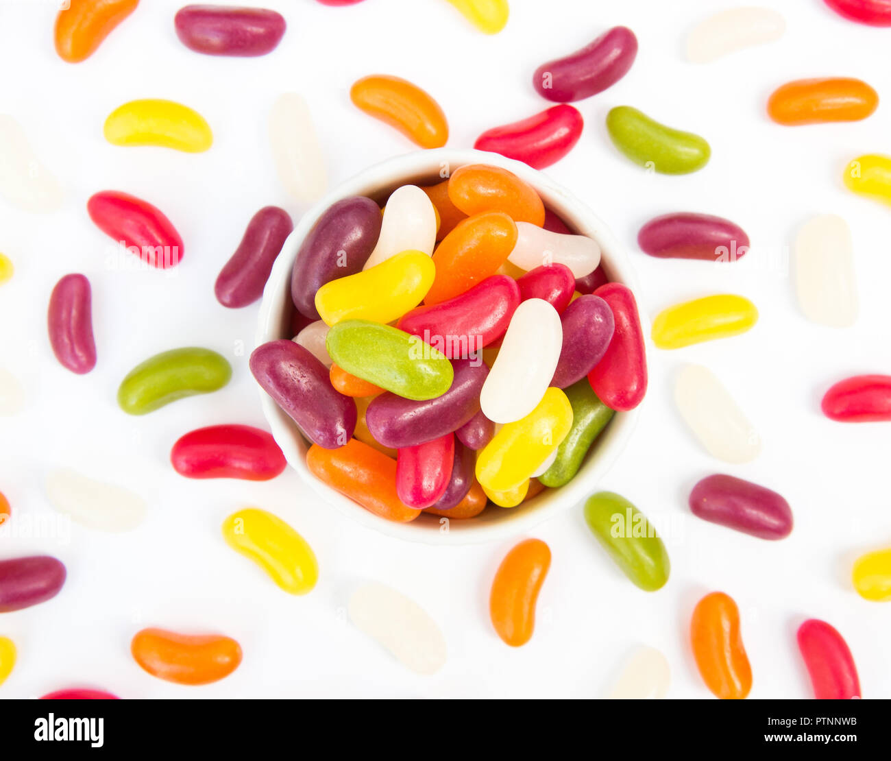 Looking down from above onto a bowl full of colourful jellybeans and candy scattered over a white background Stock Photo