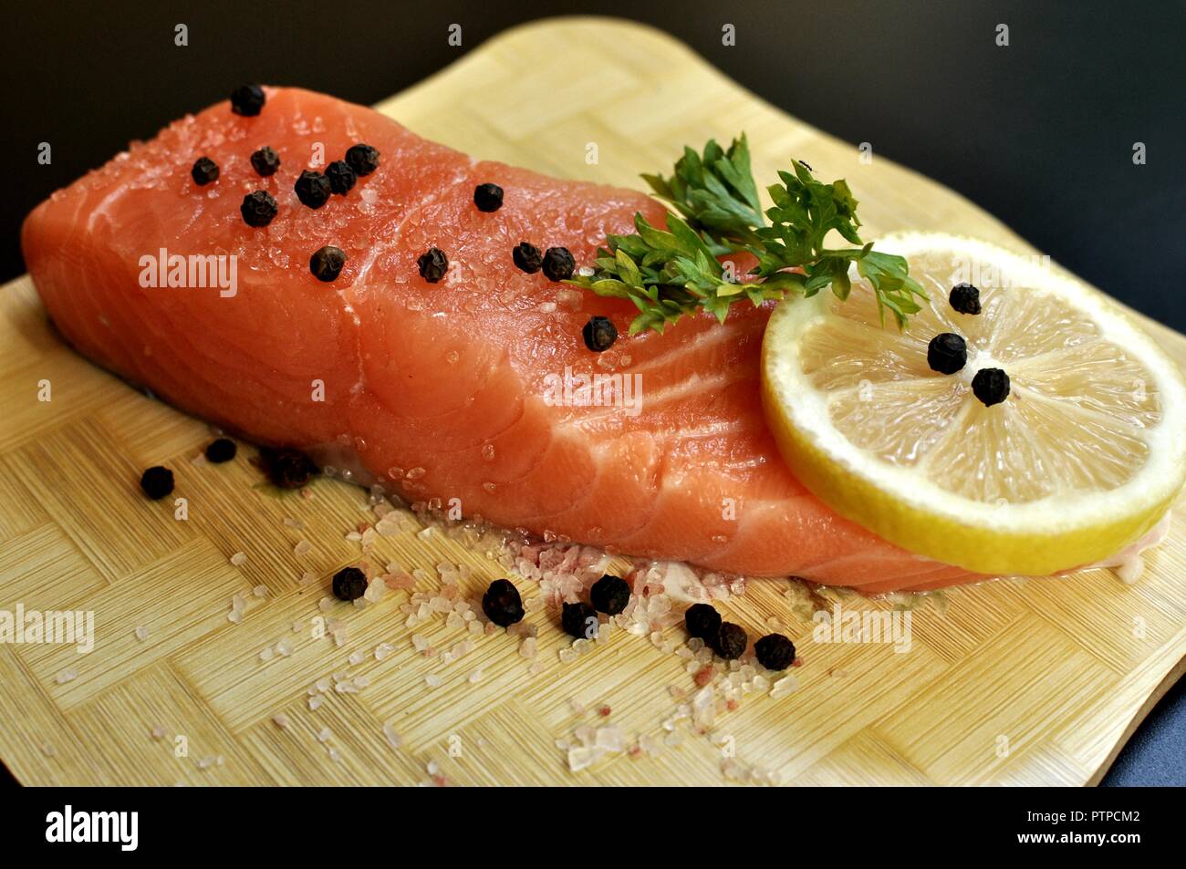 A slice of raw salmon seasoned with parsley, pepper, salt and lemon on wooden chopper and black background. Side front view Stock Photo