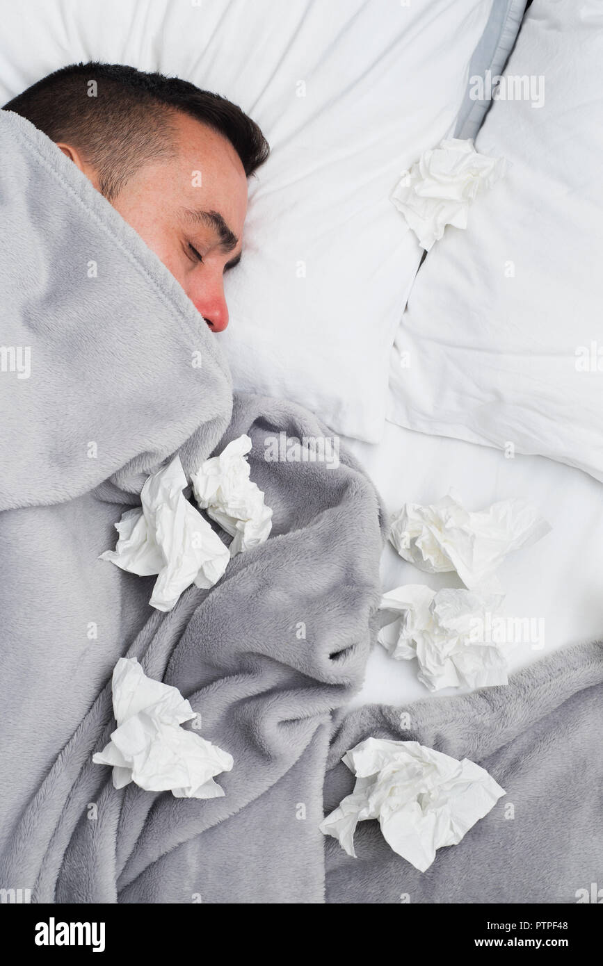 high angle view of an ill young caucasian man in bed, covered with a light gray blanket, surrounded by used tissues Stock Photo