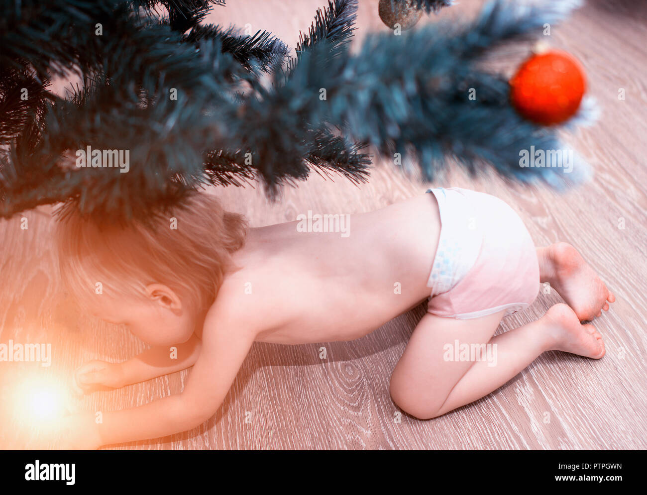 A small child in a diaper looks under the tree glowing wonderful gift, magic for the new year, fairy-tale, smile Stock Photo