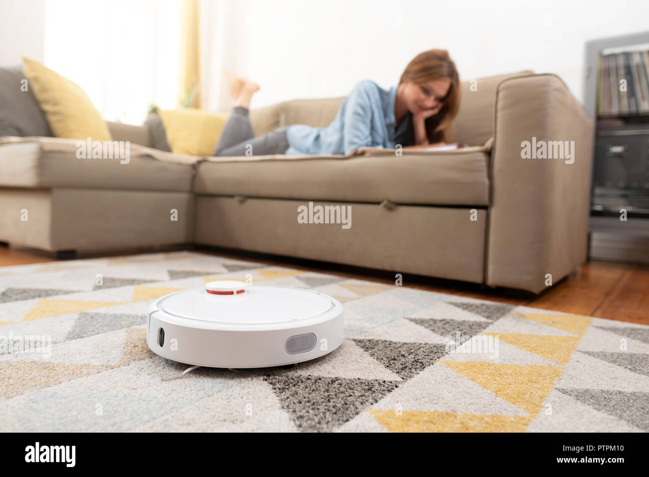Robotic vacuum cleaner cleaning the room while woman relaxing on sofa. Woman controlling vacuum with remote control. Stock Photo