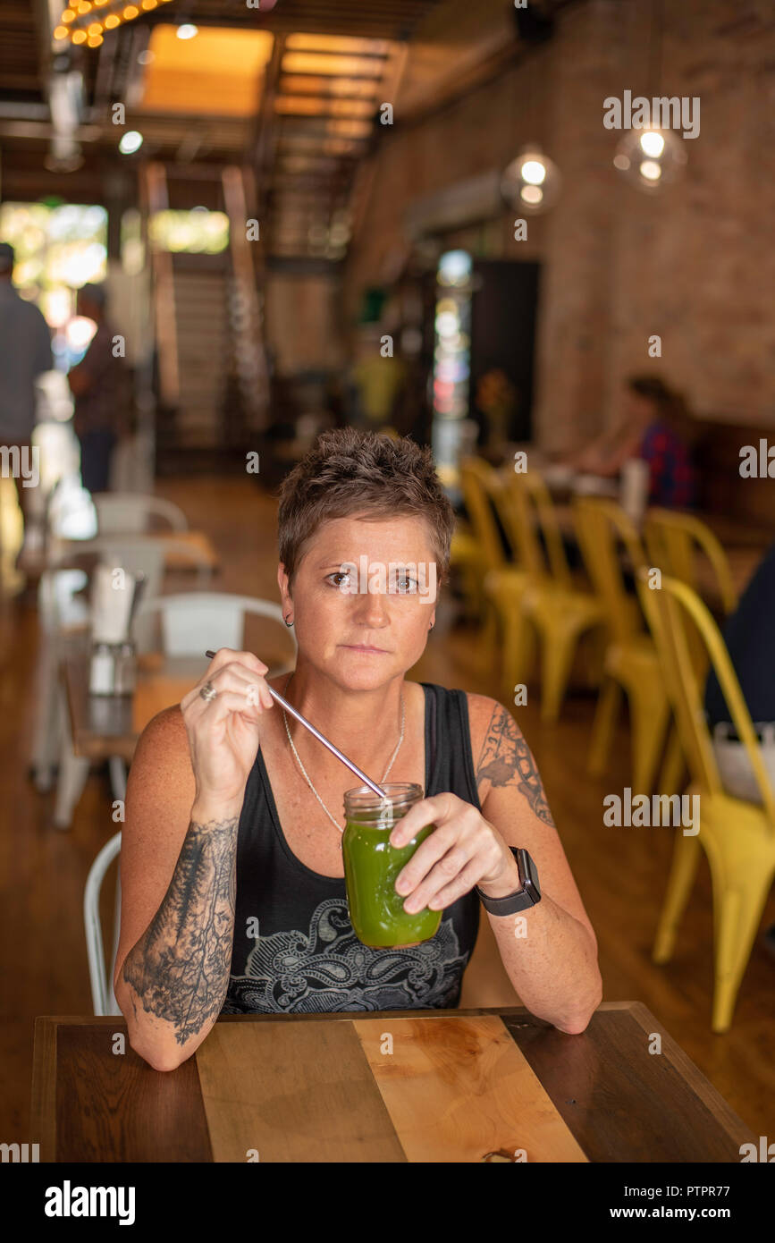 Woman with arm tattoos holding fresh pressed green juice with sustainable metal straw at the Yellow Brick Cafe in downtown Twin Falls, Idaho. Stock Photo