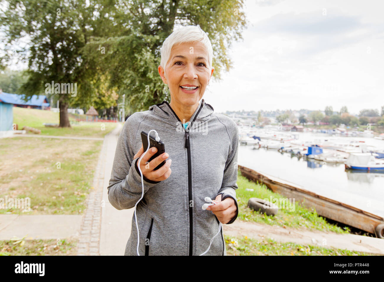 Portrait Of Active Senior Running Woman Listening Music With Smartphone In Nature, Workout Outdoor. Stock Photo