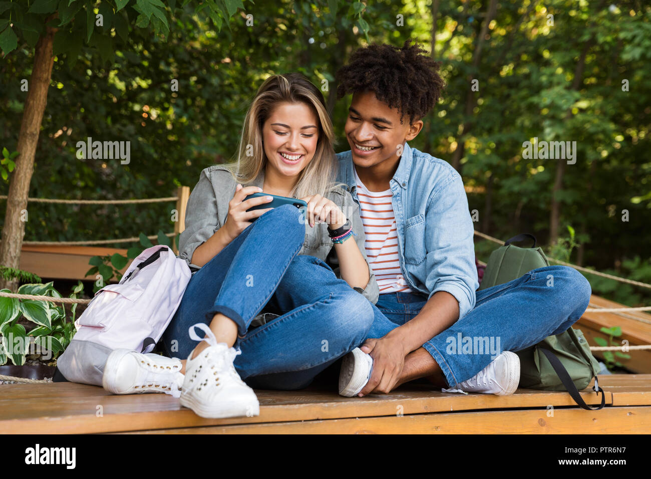 Smiling young multiethnic couple talking while spending time together at the park, using mobile phone Stock Photo