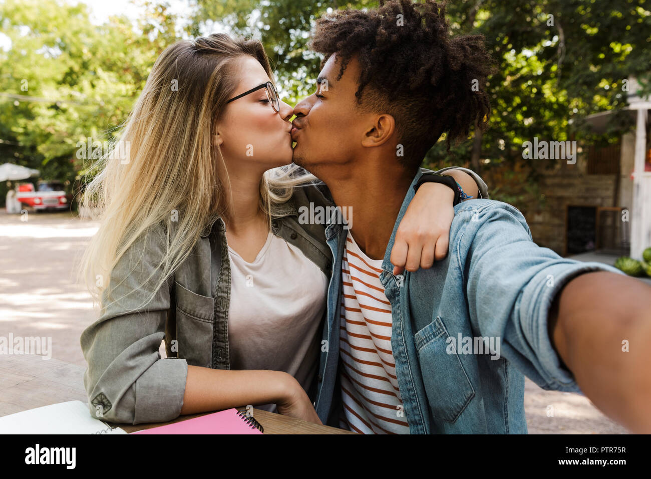 Kissing young multiethnic couple spending time together at the park, studying while sitting at the table and taking a selfie Stock Photo