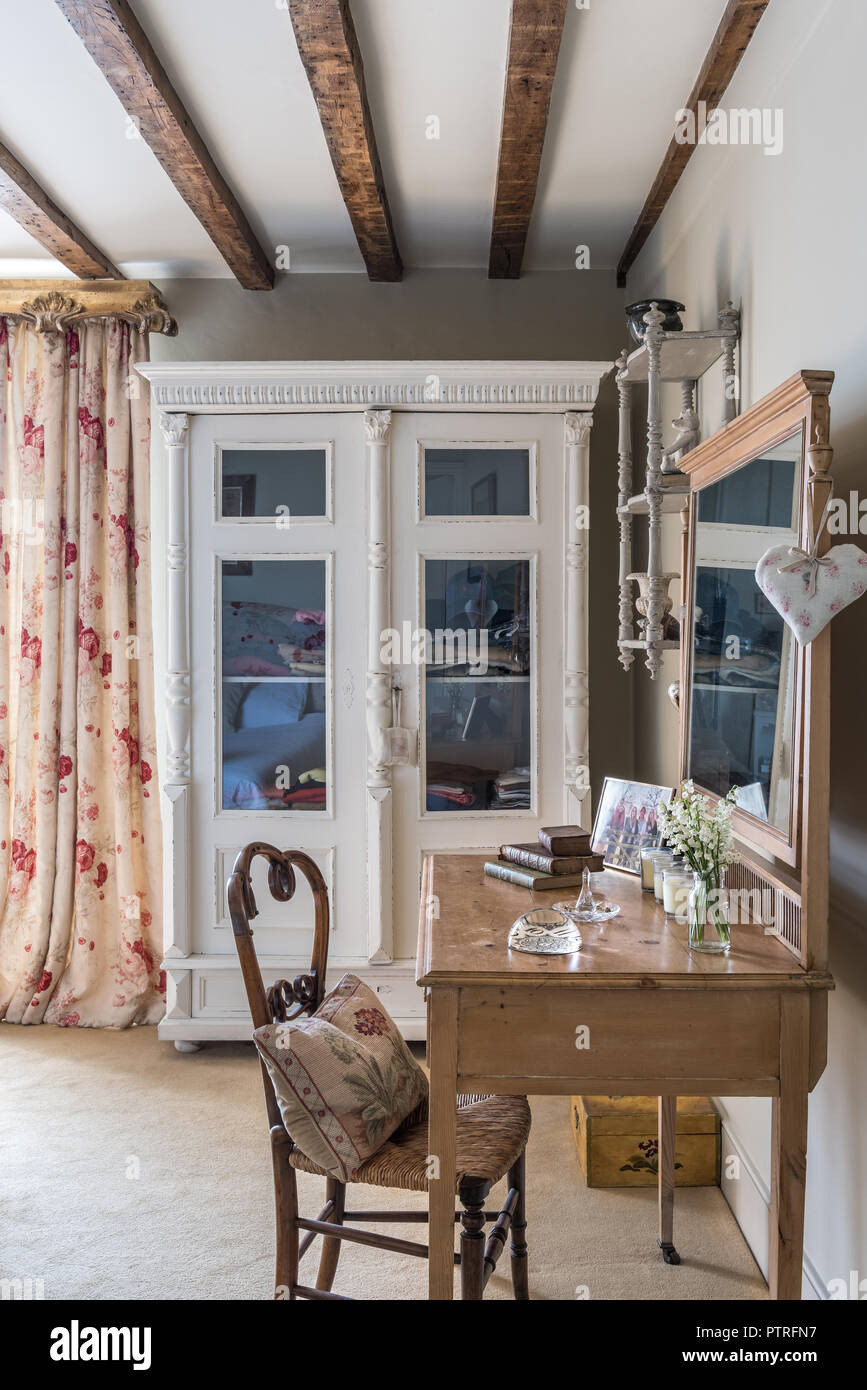 Glass fronted cabinet with floral curtains and wooden dressing table with wicker chair in 16th century farmhouse renovation Stock Photo