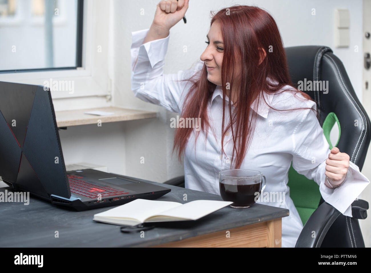 a young woman at a desk with a laptop raises her hand up in the gesture of success, behaves as if she's gonna win Stock Photo