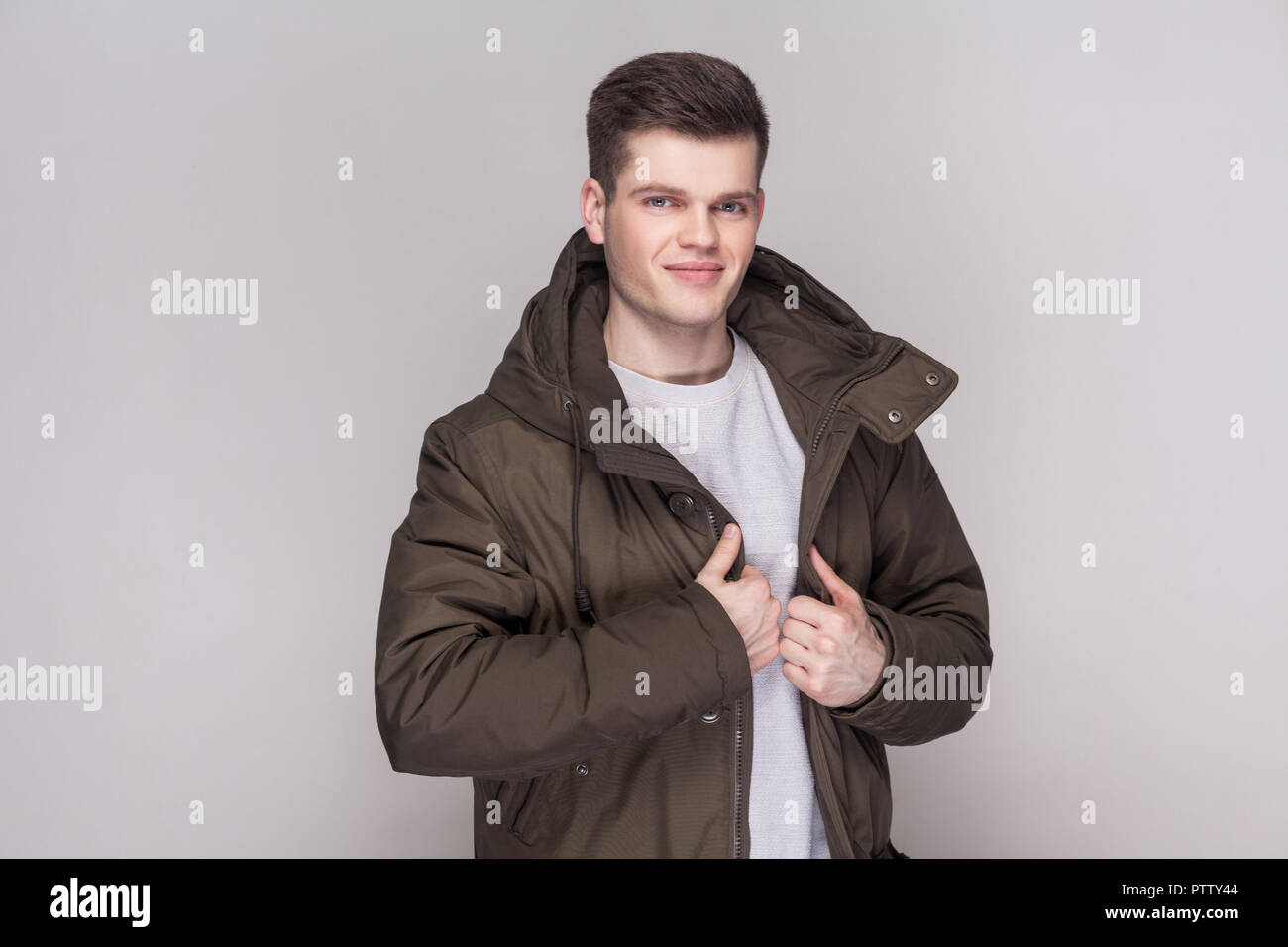 Portrait of young handsome man standing in light gray shirt and dark green parka looking at camera with smile and satisfied face. indoor studio shot,  Stock Photo