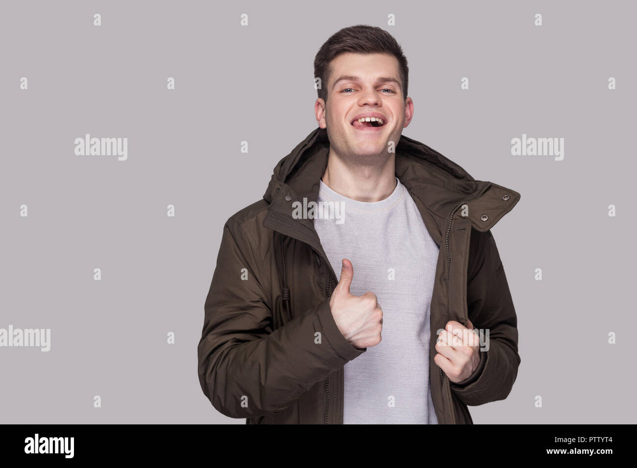 Portrait of young funny handsome man standing in light gray shirt and dark green parka looking at camera and showing thumbs up. indoor studio shot, is Stock Photo