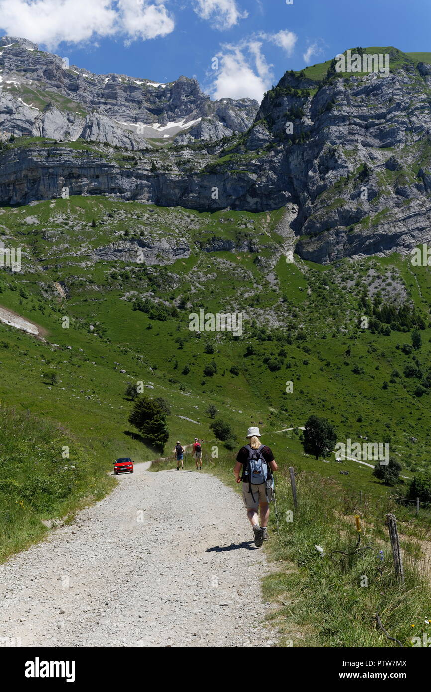 Female hiker walking on the off road tracks with hikers and red car in the distants in the valleys of the french alps nr Montmin France Stock Photo
