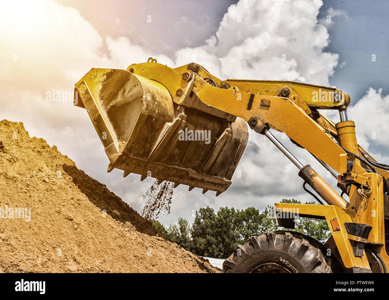 Yellow forklift lifted bucket on a pile with earth, blue sky and sun Stock Photo
