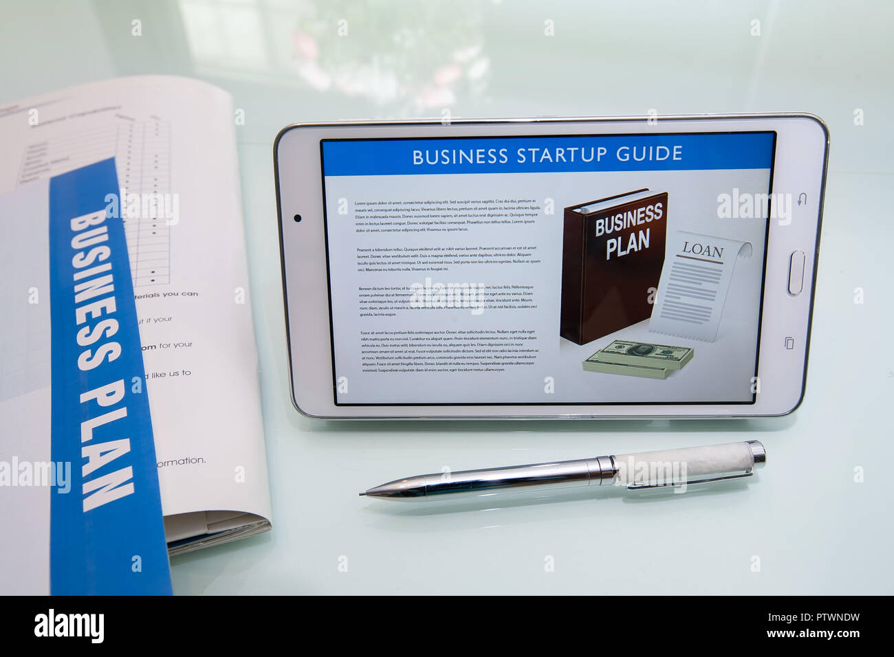 Writing a business plan using a business startup guide, printed collateral, pen, and tablet or mobile device on a reflective glass desk. Stock Photo