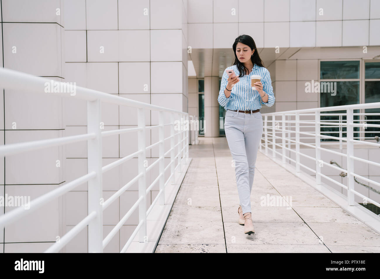 a full length photo of an elegant lady using phone while walking through the bridge, she is holding a cup of coffee Stock Photo