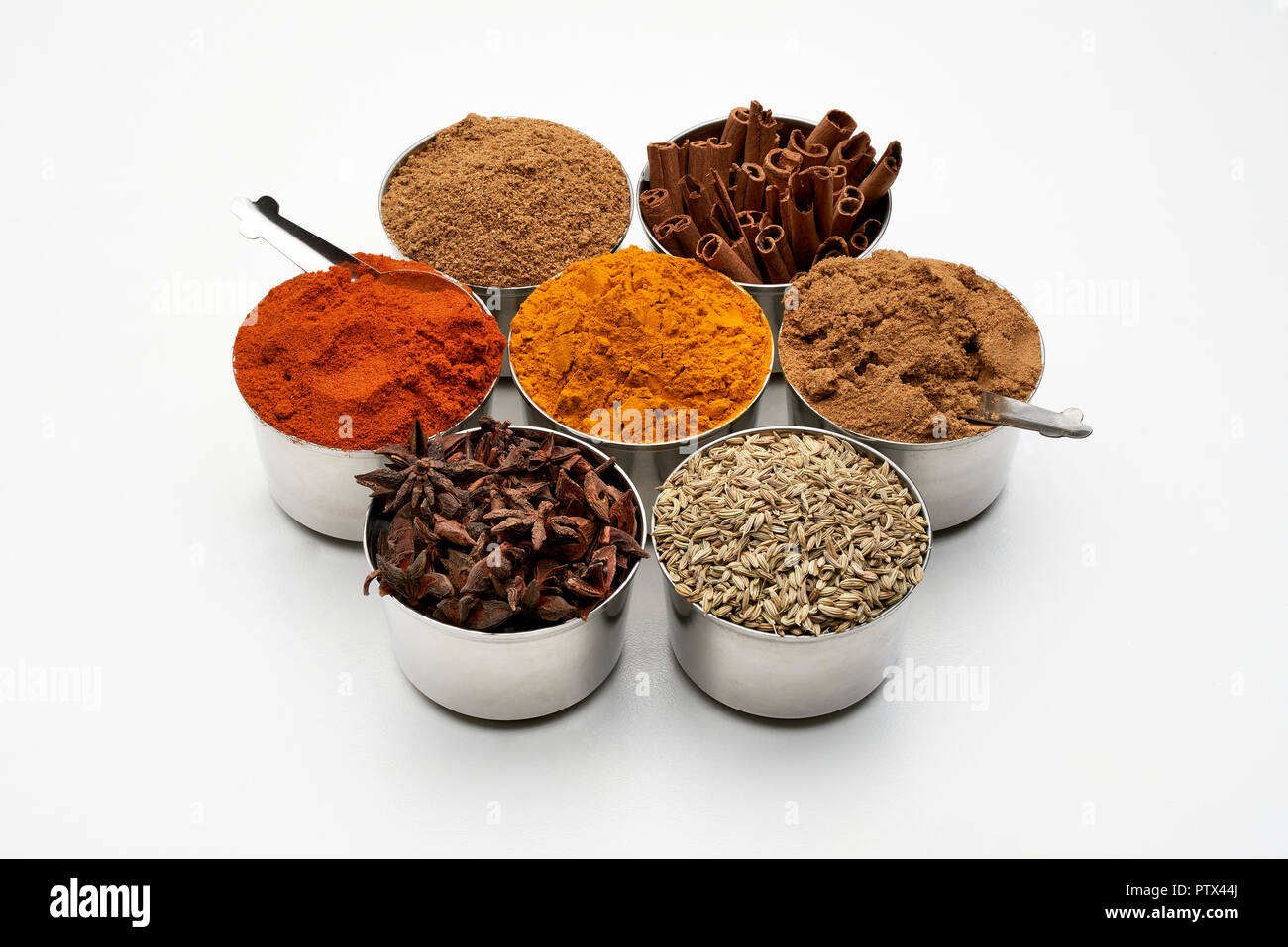 Seven spice tins on a white background including, chili, turmeric, garam masala and cumin powders as well as cassia bark, star anise and fennel seeds. Stock Photo