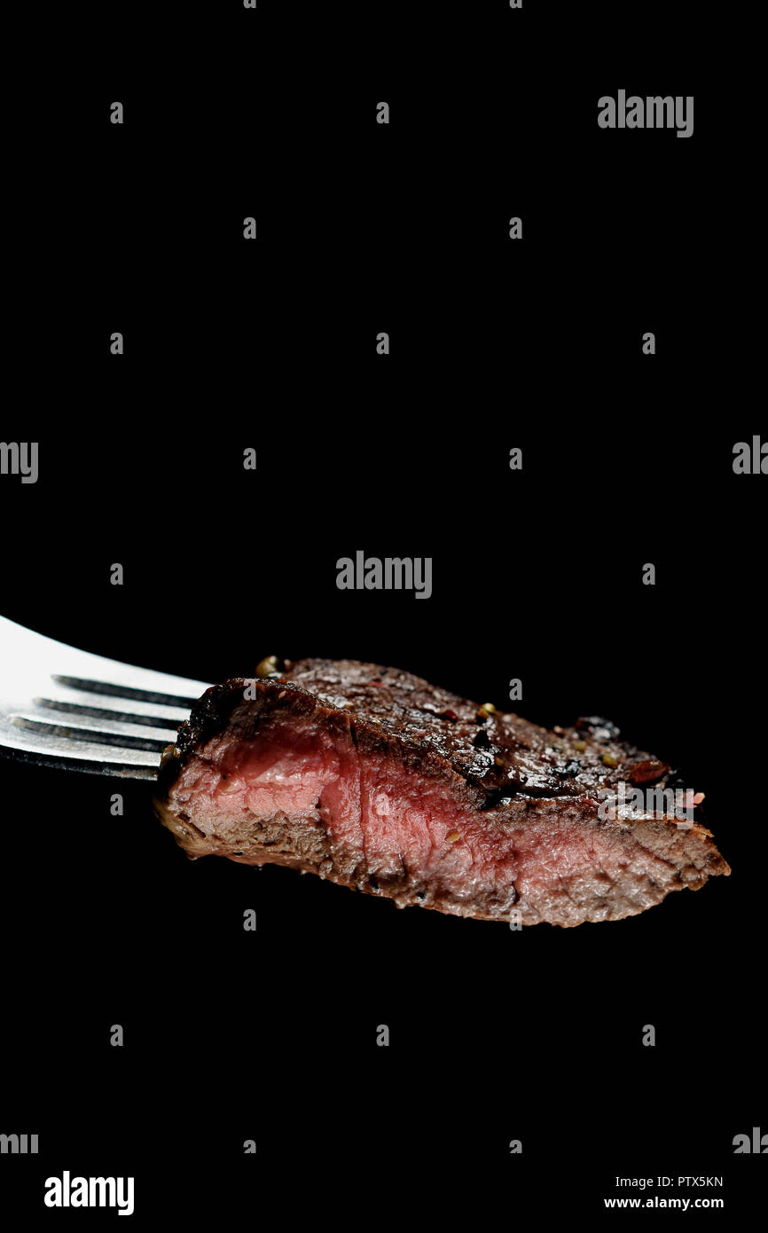 Piece of a grilled medium rare steak on a fork. Vertical image with copy space. Isolated on black. Stock Photo