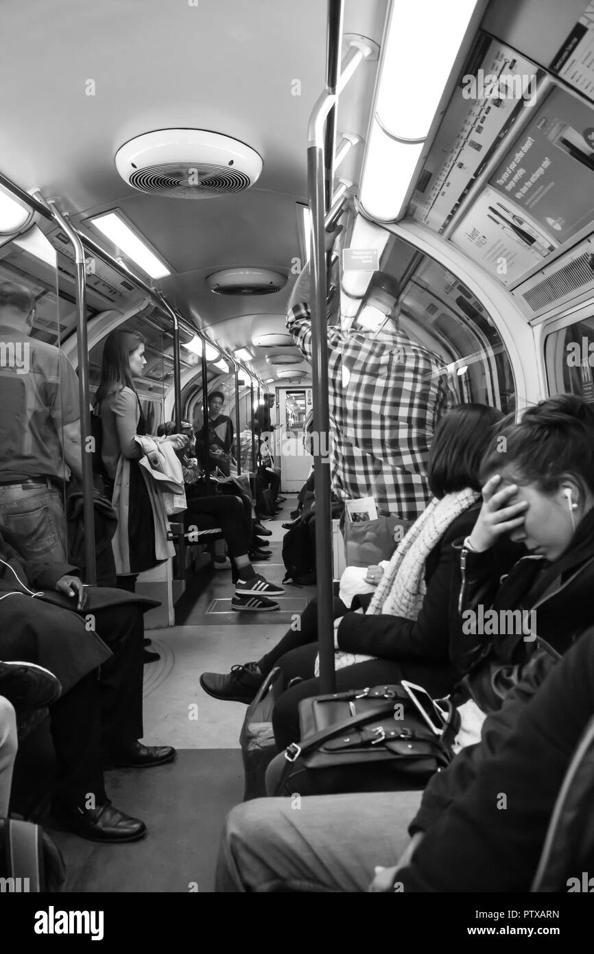 Black & white interior view of tube train passengers; commuters standing & sitting. Anxious young adult holds head in hands, face palming gesture. Stock Photo