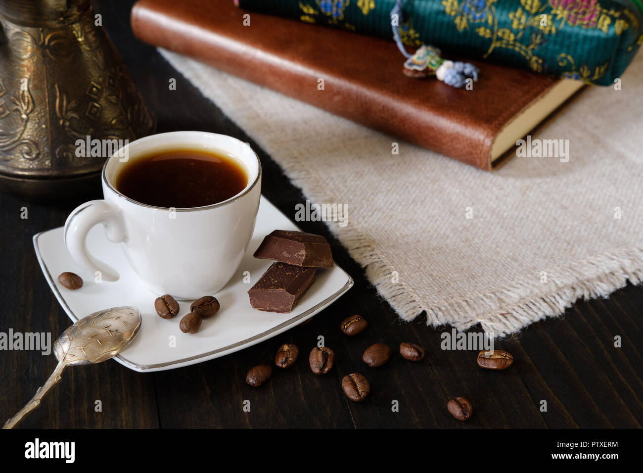 Small cup of black coffee with vintage teaspoon, turkish coffee pot and chocolate pieces on dark wooden background with diary and pencil case Stock Photo