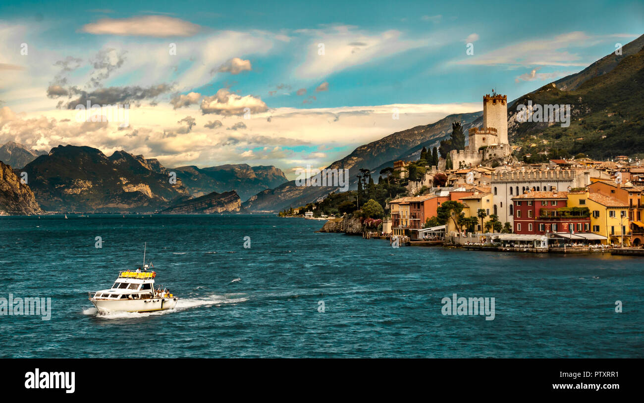 View of Lake Garda with white clouds over blue skies, a boat in the foreground and the colourful town of Tignale and mountains in the background Stock Photo