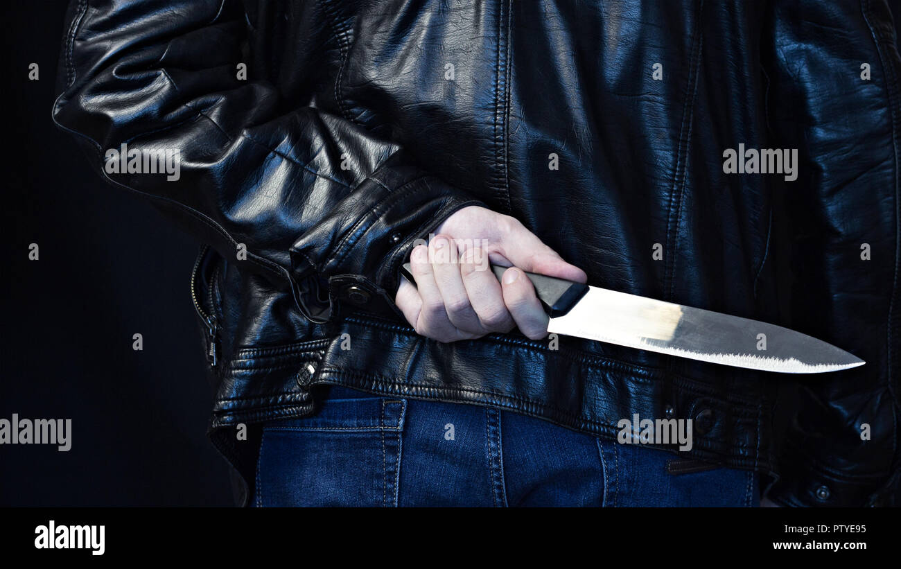 A man in a leather jacket holds a knife behind his back, a black background Stock Photo