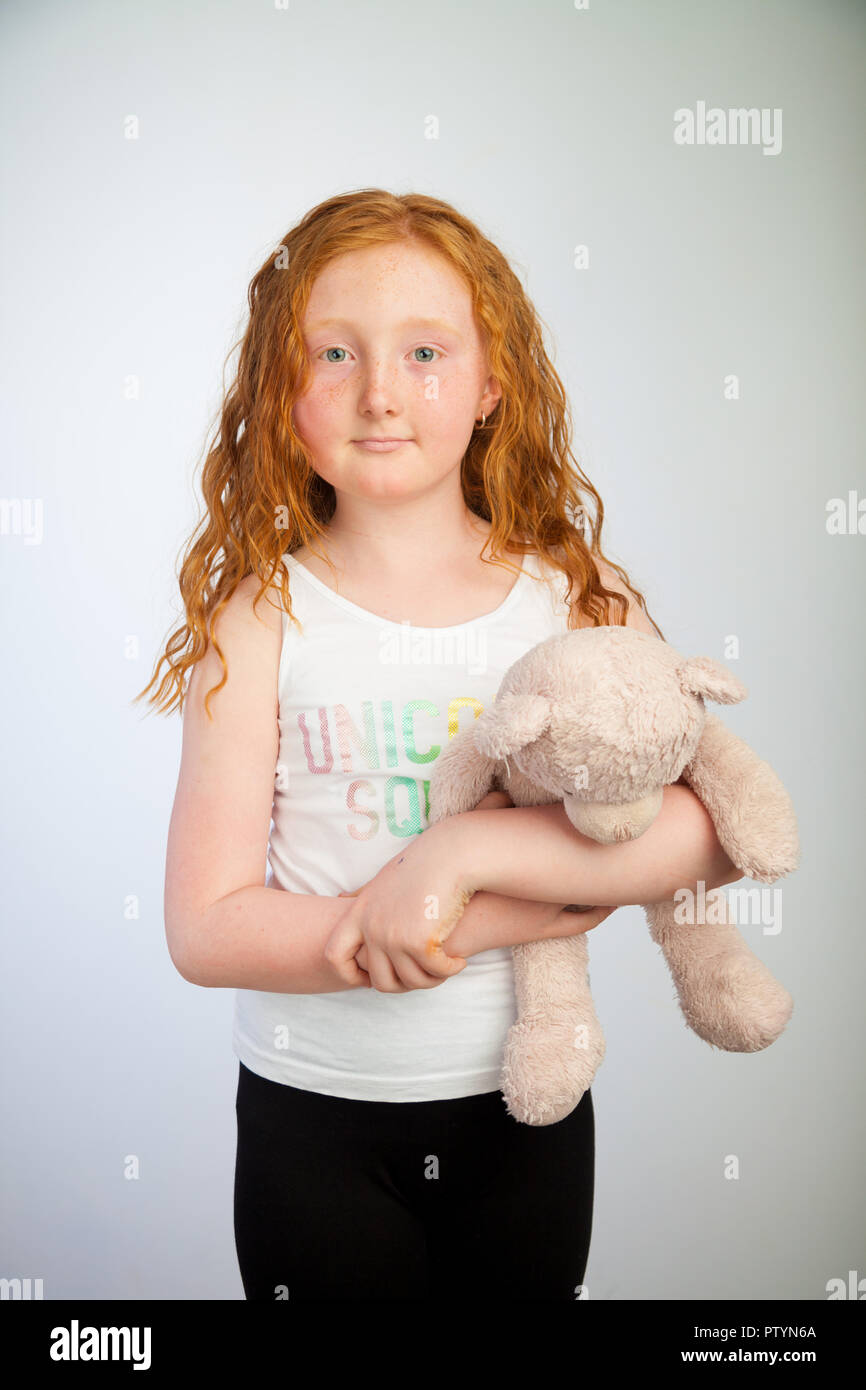 Portrait of a 10 year old girl with ginger hair and freckles standing holding a cuddly toy. Stock Photo