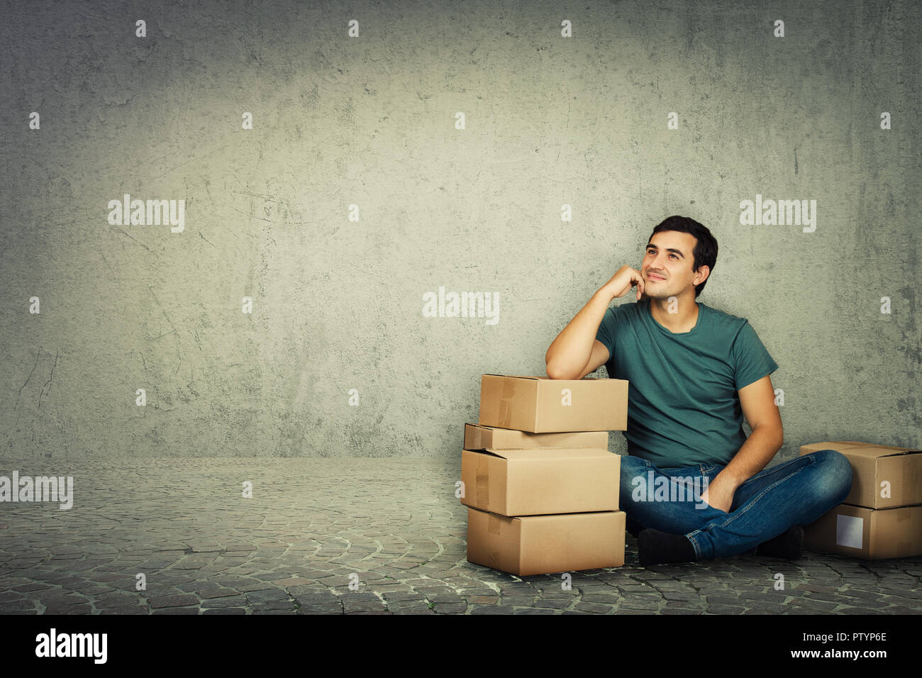 Pensive young man sitting and leaning on cardboard boxes hand on cheek and dream. Ready for moving to a new house over grey wall background. Stock Photo