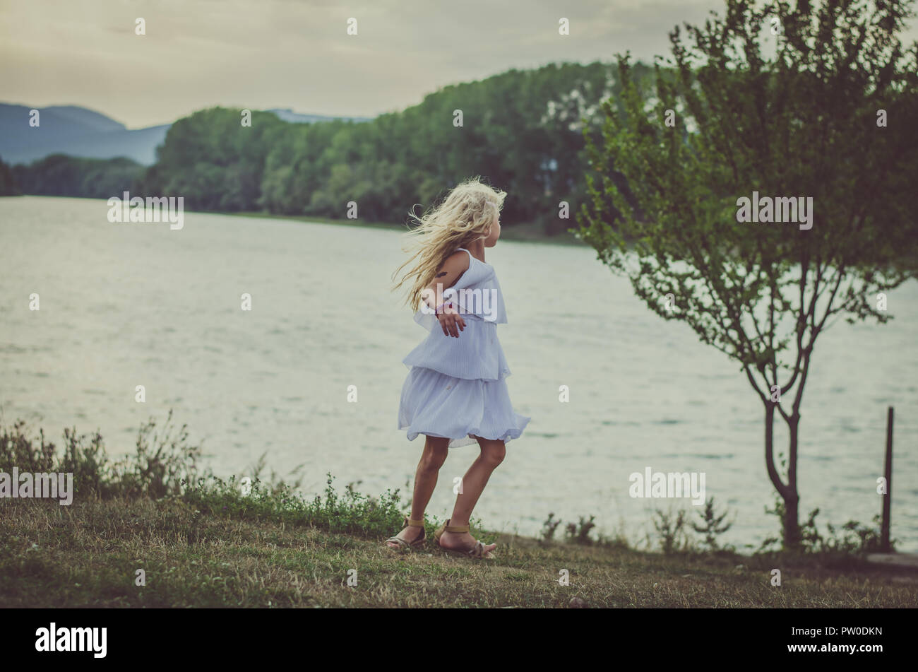 adorable little girl in dress at the pond outdoors in the afternoon having fun  at orange sunset back view Stock Photo