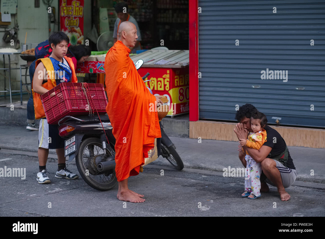 A man and his little daughter in a market in Phuket Town, Thailand, inviting the blessing of a monk, the monk's helper behind to help carry alms Stock Photo