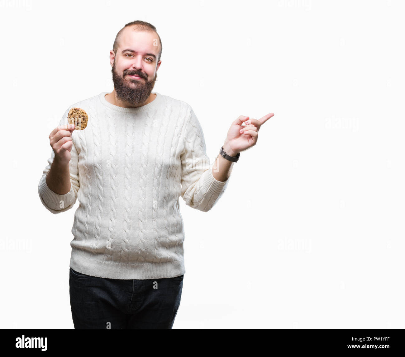 Young hipster man eating chocolate chips cookie over isolated background very happy pointing with hand and finger to the side Stock Photo