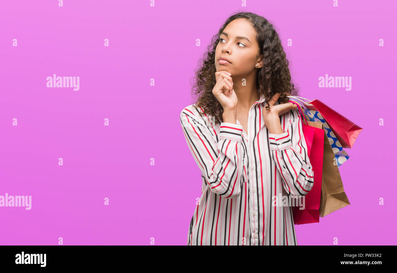 Young hispanic woman holding shopping bags serious face thinking about ...