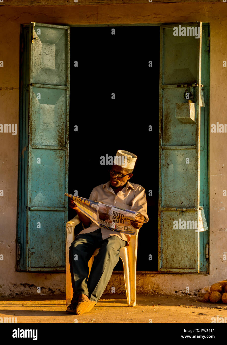 An Old Man with a newspaper sitting in front of his shop Stock Photo