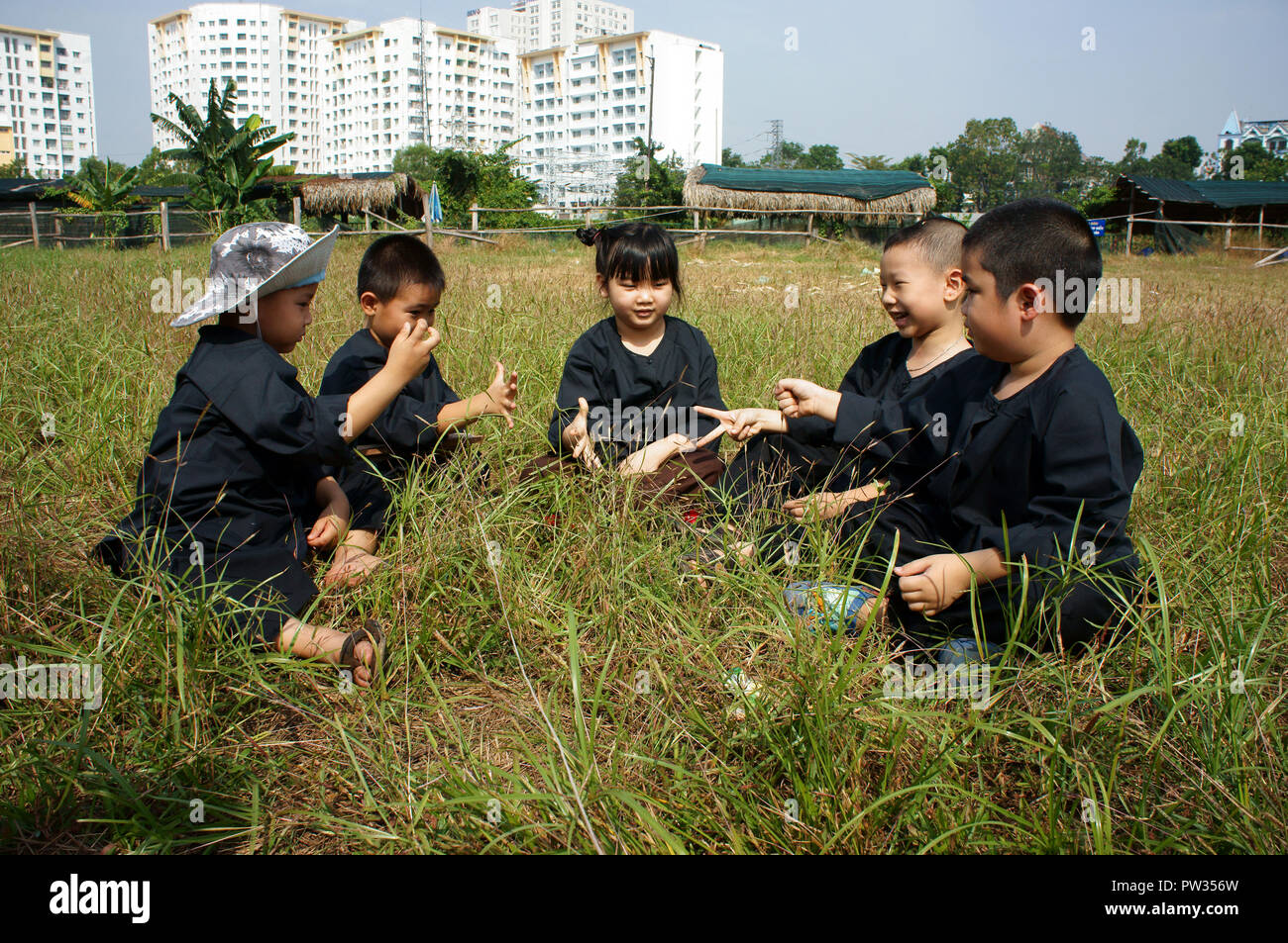 HO CHI MINH CITY, VIET NAM, Group of Vietnamese children in black traditional dress, sit on grassland and play together, outdoor activity Stock Photo