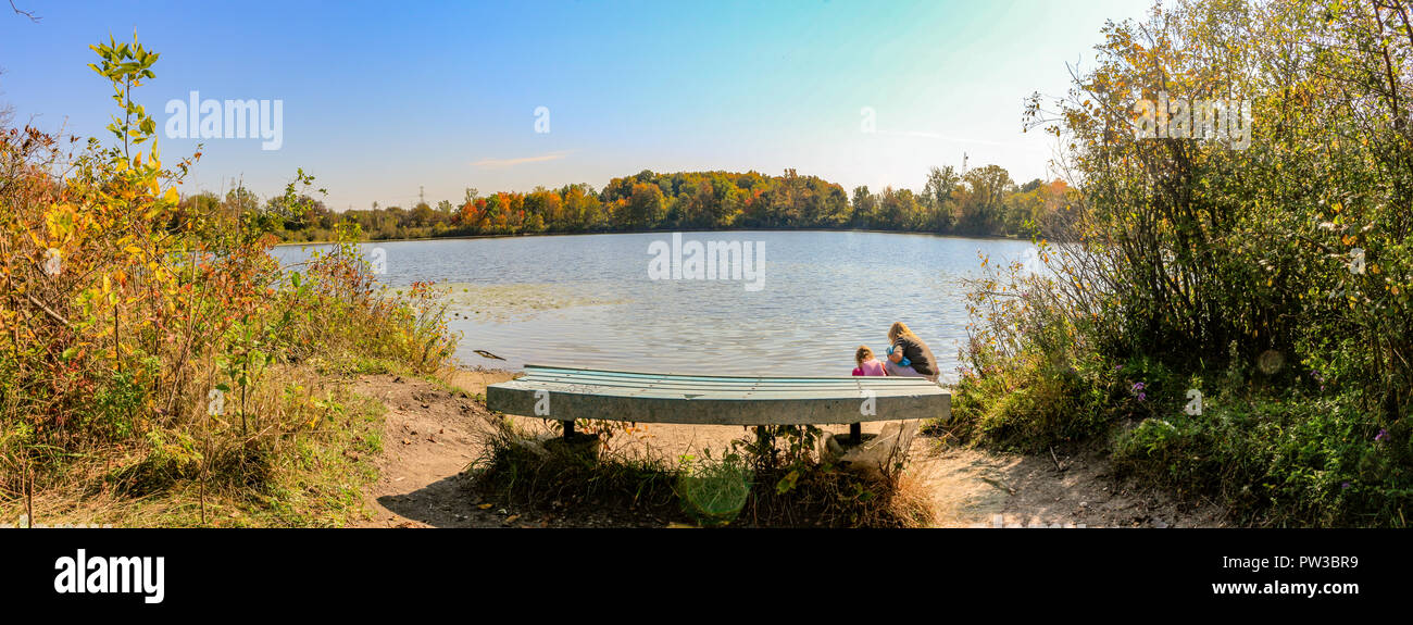 Panorama format landscape photo of a bench overlooking a lake with 2 people looking at the water. Beautiful Canadian fall weather. Stock Photo