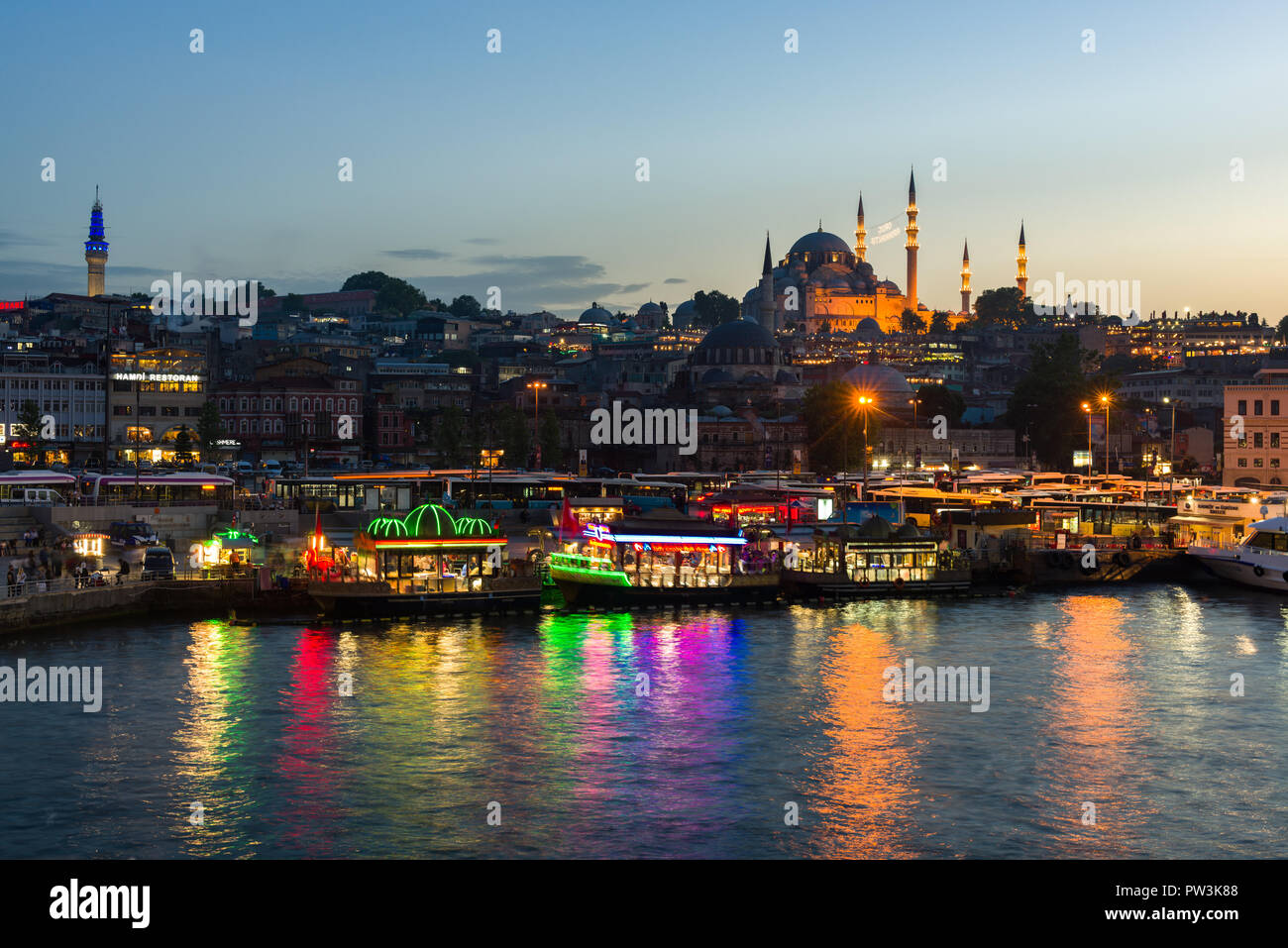 The brightly lit floating kitchen boats of Eminonu with Suleymaniye mosque and Golden Horn in background at dusk, Istanbul, Turkey Stock Photo