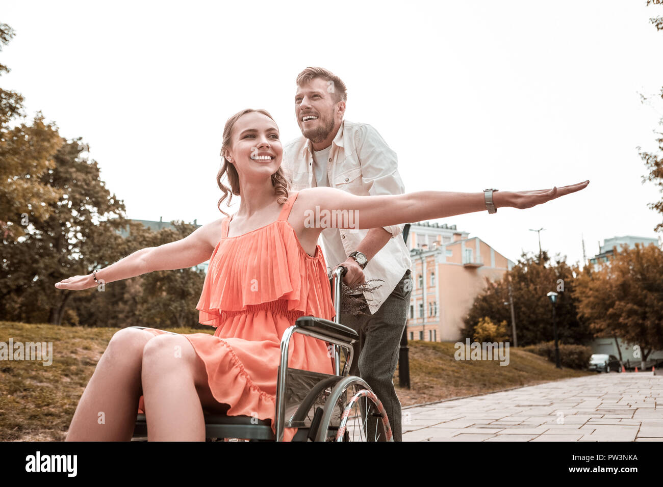 Reliable man smiling and pushing the wheelchair of his happy girlfriend Stock Photo