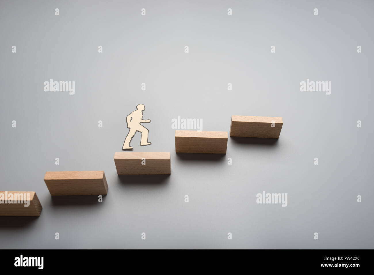 Paper cutout of a businessman walking up the ladder of success made of wooden blocks over grey background. Stock Photo