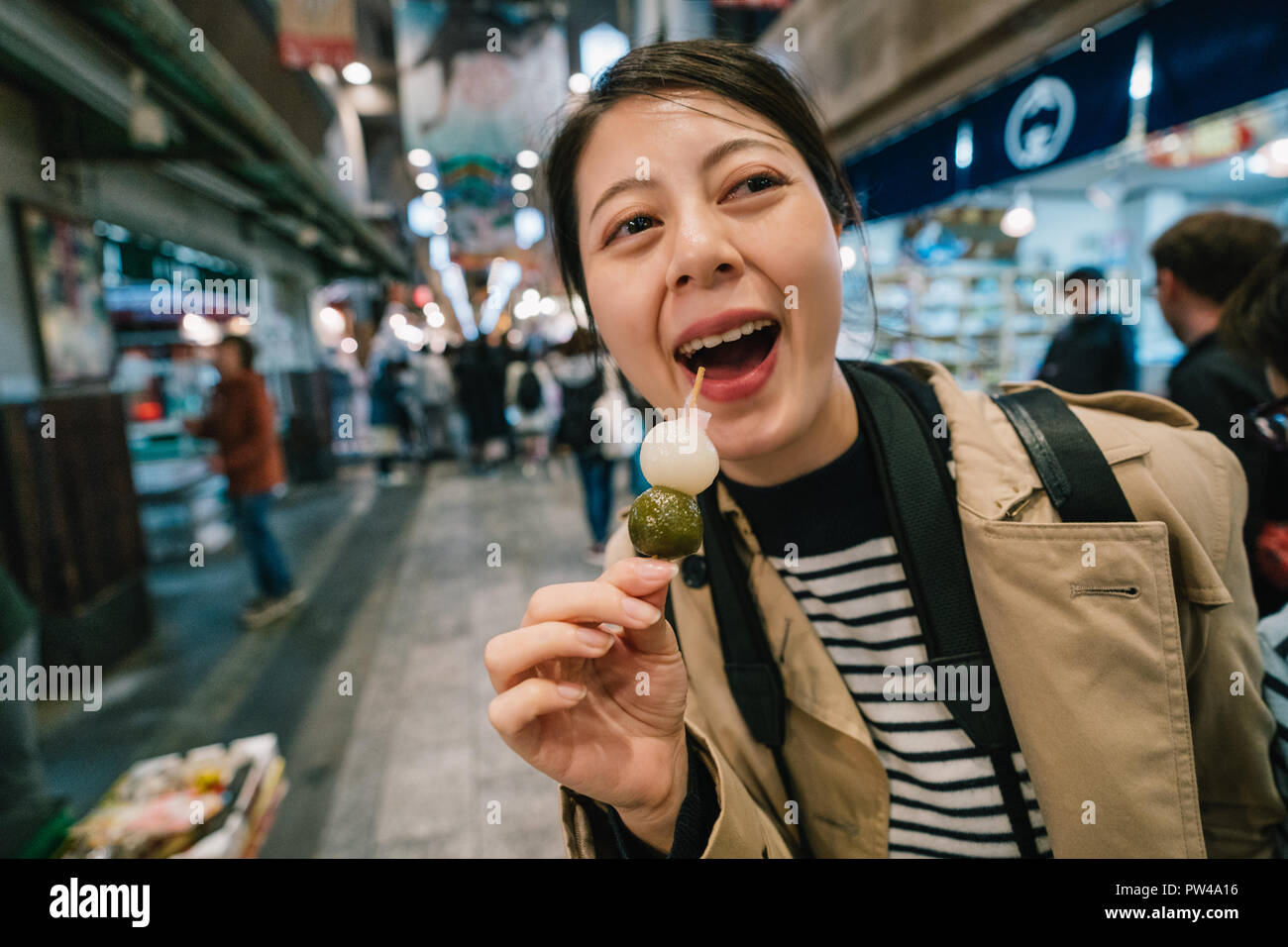 close up photo of an elegant female traveler joyfully trying the Japanese street food. Cheerful tourist shopping at outdoor traditional market in Kyot Stock Photo