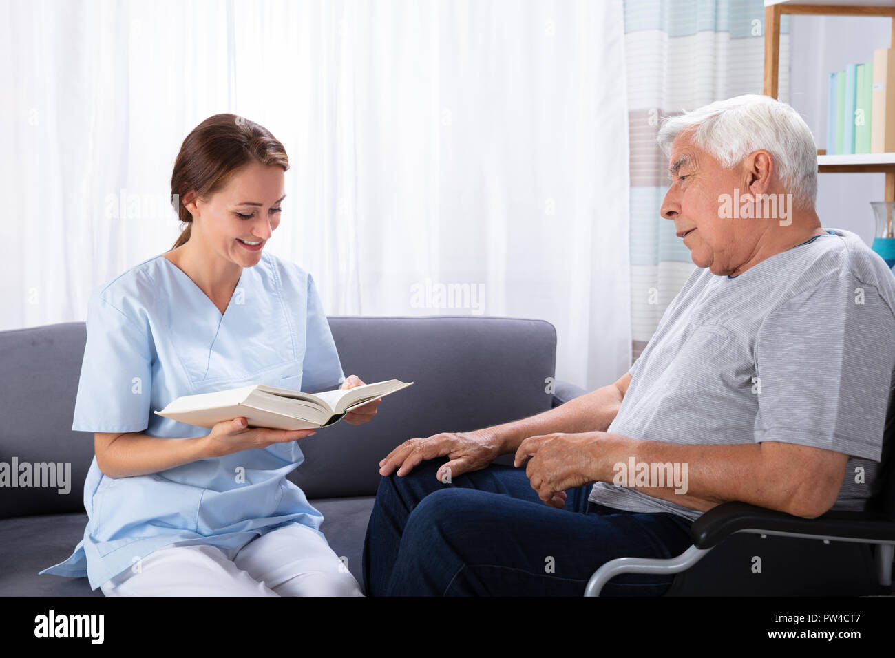 Nurse Reading Book For Disabled Senior Man Sitting On Wheelchair Stock Photo