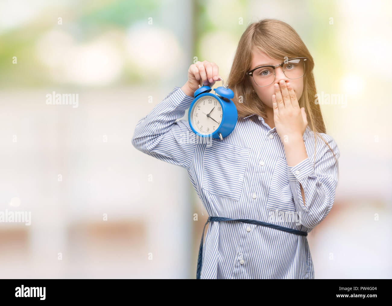 Young blonde child holding alarm clock cover mouth with hand shocked