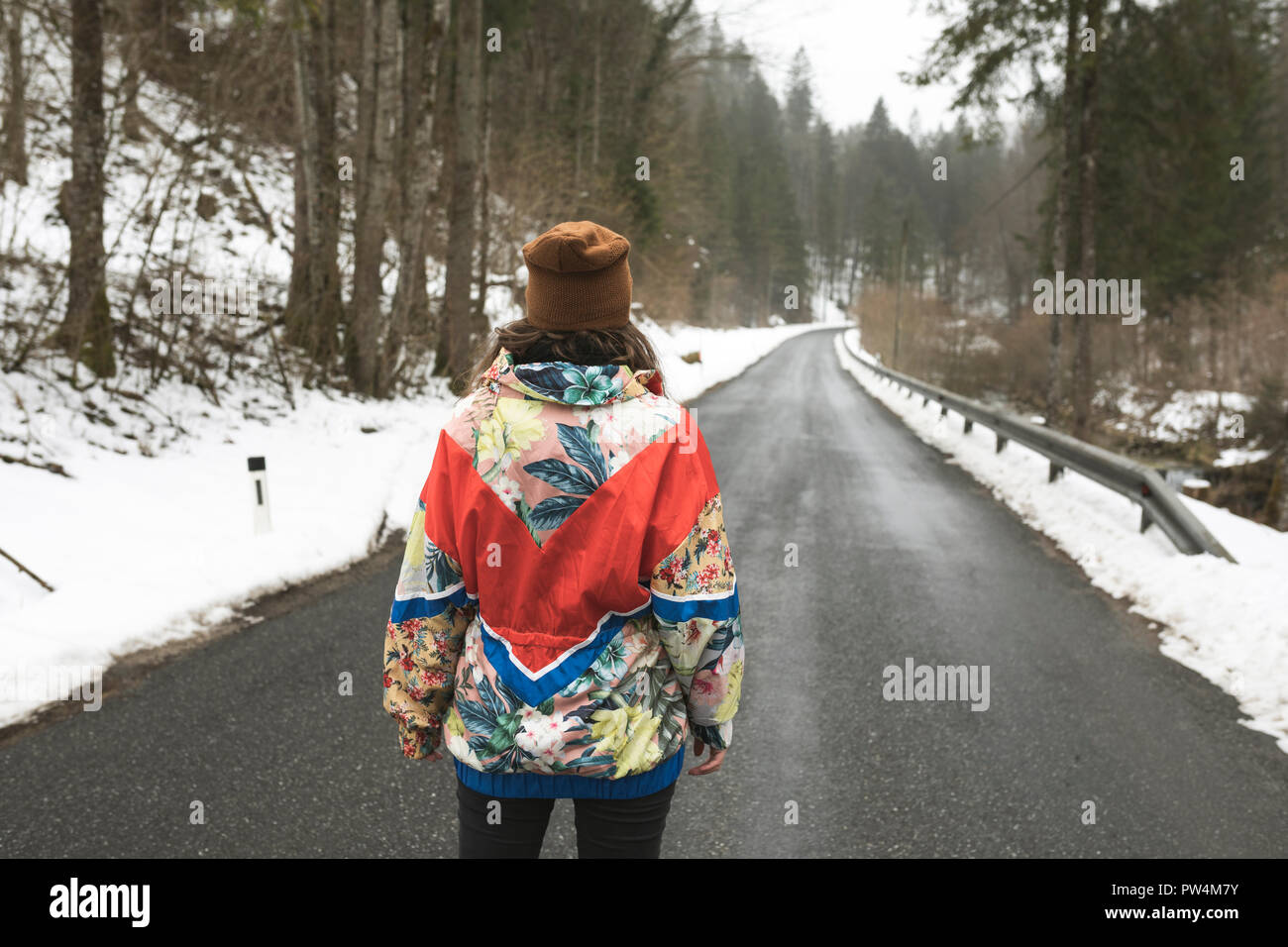 Rear view of woman wearing warm clothing standing on country road amidst trees in forest during winter Stock Photo