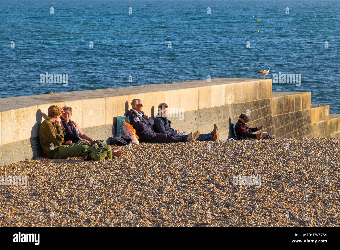 People sitting on the beach enjoying the late evening sunlight in the coastal town of Lyme Regis in Dorset. Stock Photo