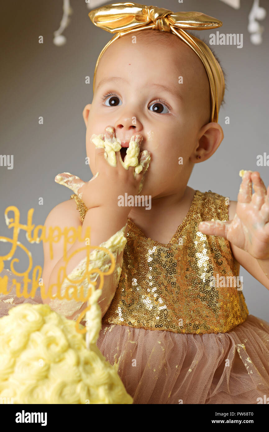 small sweet baby girl in a golden dress with a bow on her head trying a jazzy jelly cake from a cream. studio shot of a birthday on a gray background surrounded by balls Stock Photo