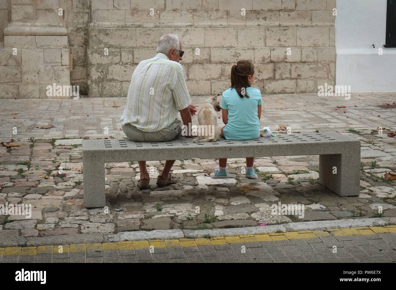 A grandfather and his granddaughter sit with their pet dog outside the Parroquia De Nuestra Señora Del Rosario, in Es Castell, Menorca, Spain Stock Photo