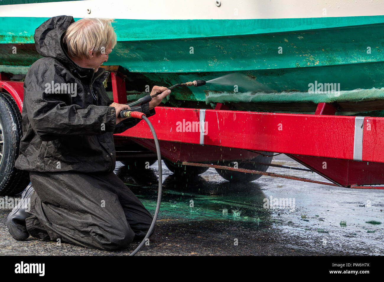 Caucasian man kneeling while high pressure washing dirty boat hull on red trailer Stock Photo