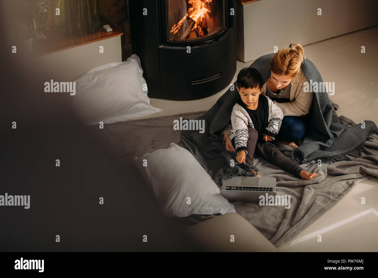 Young family sitting near fireplace looking at laptop. Son pointing at laptop to show something interesting to his mother at home. Stock Photo
