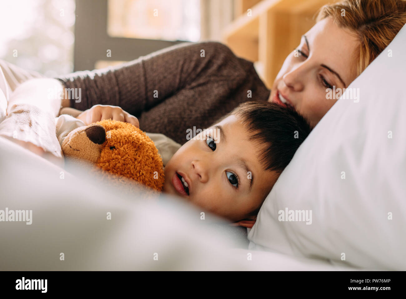 Cute little boy holding his teddy bear and lying on bed with his mother. Small kid in bed with his toy and mother. Stock Photo