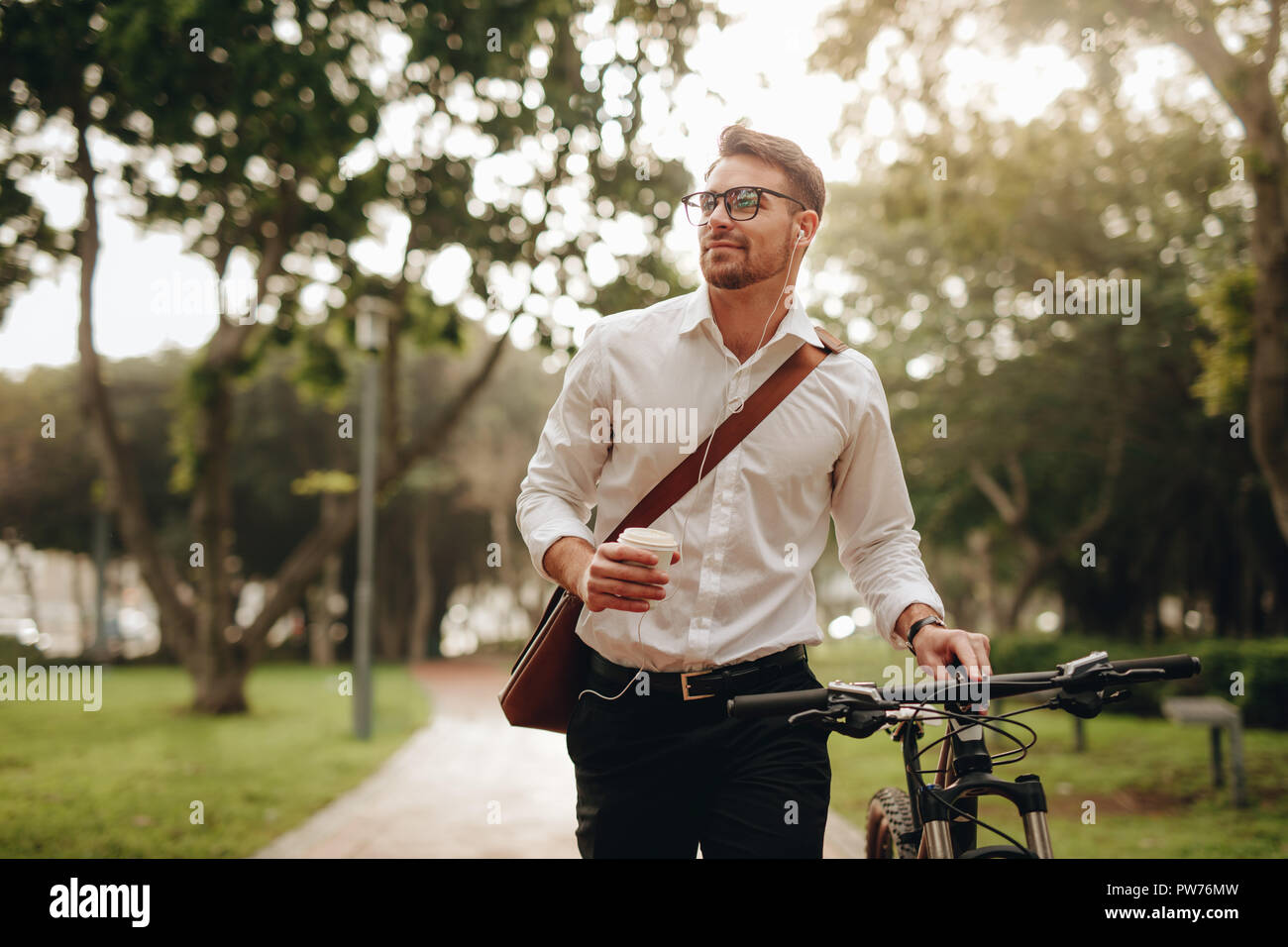 Businessman listening to music and enjoying coffee while walking to office with his bike. Man enjoying his commute to office walking with his bicycle Stock Photo