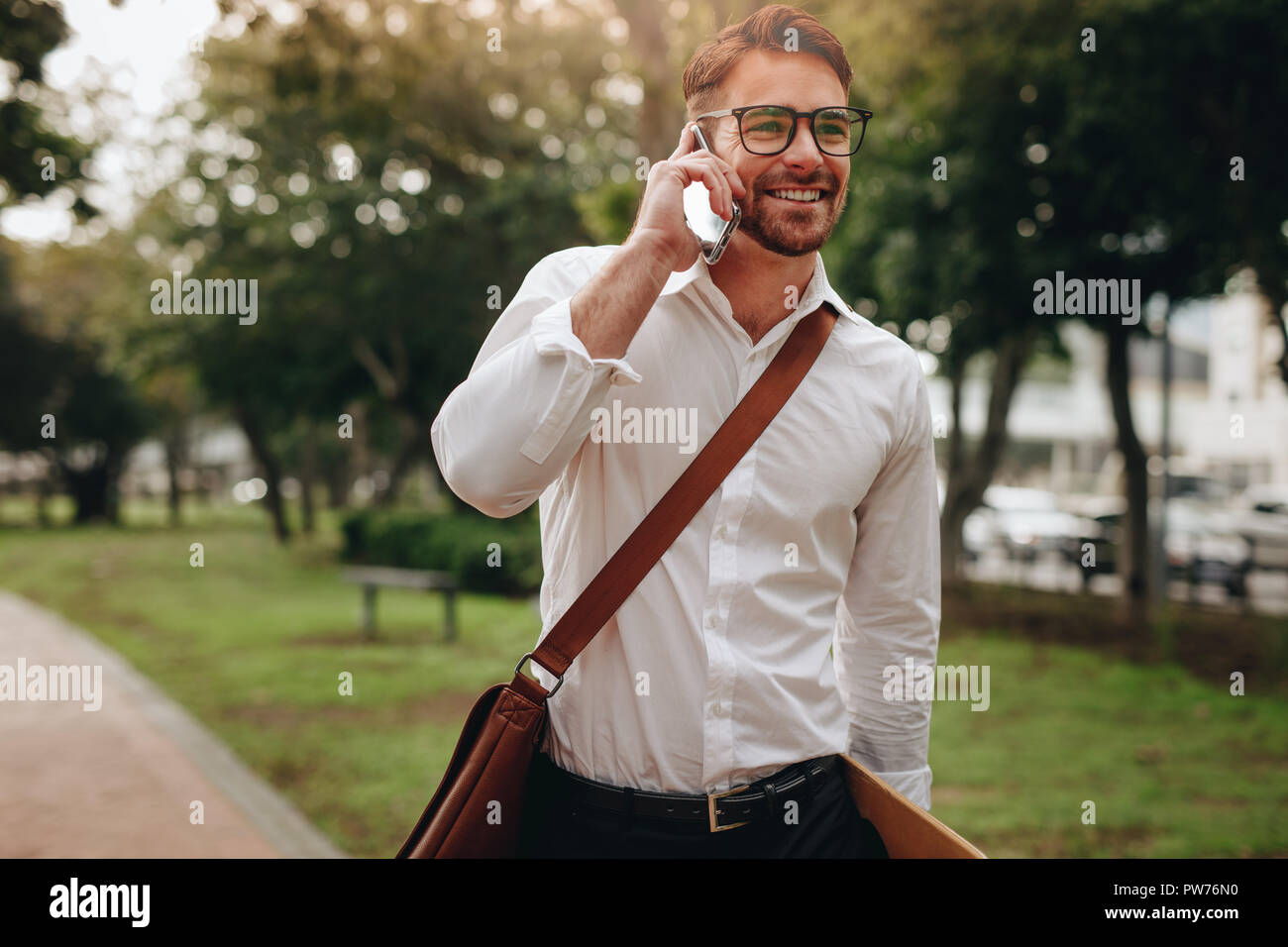 Man carrying office bag talking over mobile phone while walking to office. Smiling businessman going to office by walk early in the morning. Stock Photo