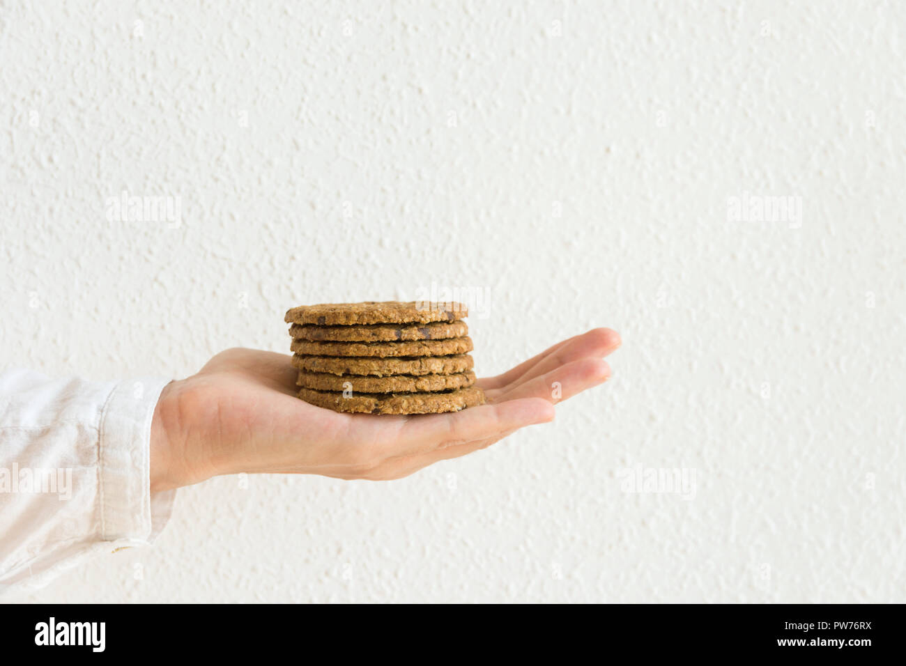 Young caucasian woman holds in stretched hand stack of chocolate chip cookies with oats bran nuts on white wall background. Holiday baking healthy pas Stock Photo