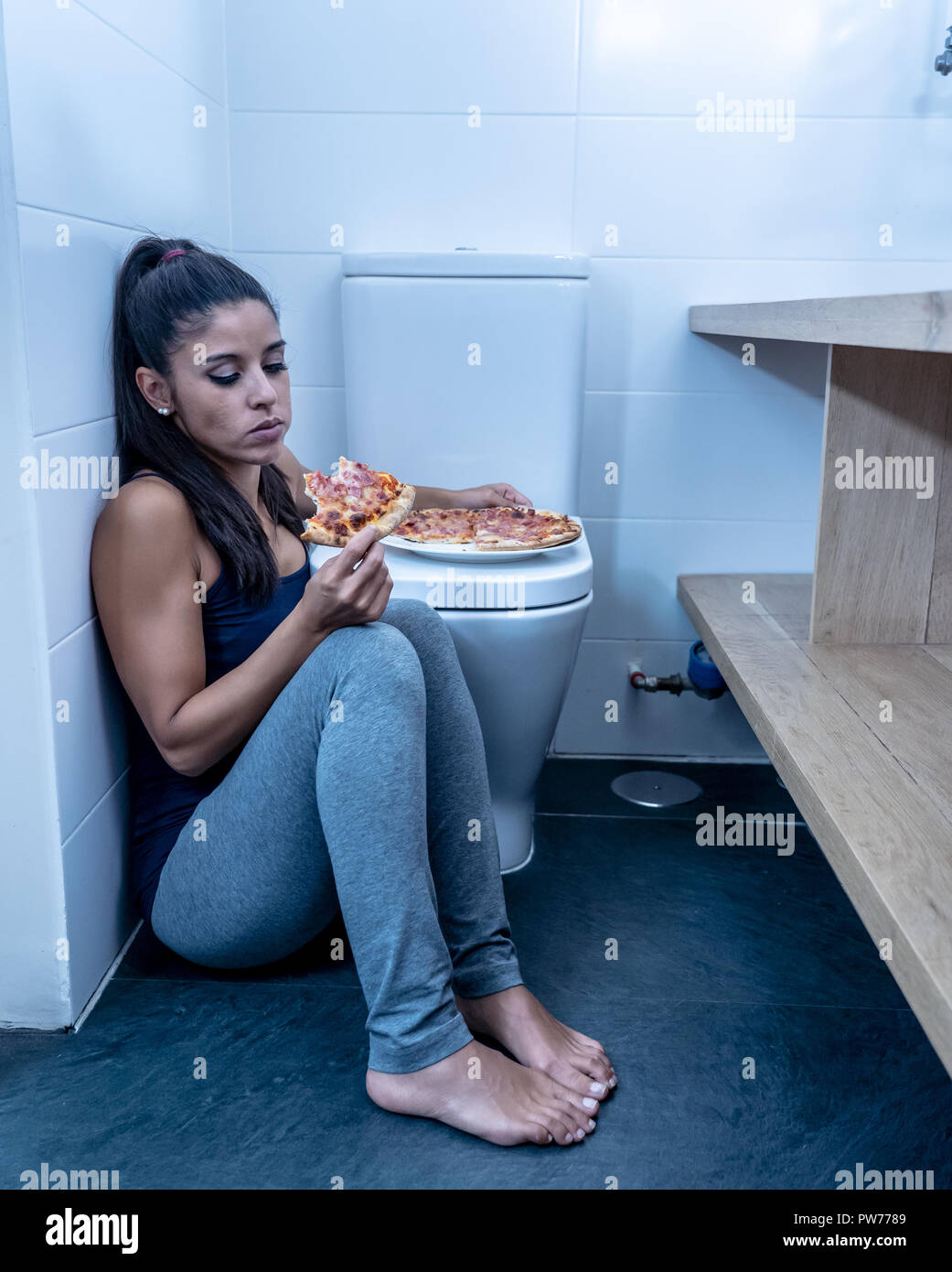 Attractive young and sad bulimic young woman feeling guilty and sick eating while sitting on the floor next to the toilet in eating disorders anorexia Stock Photo